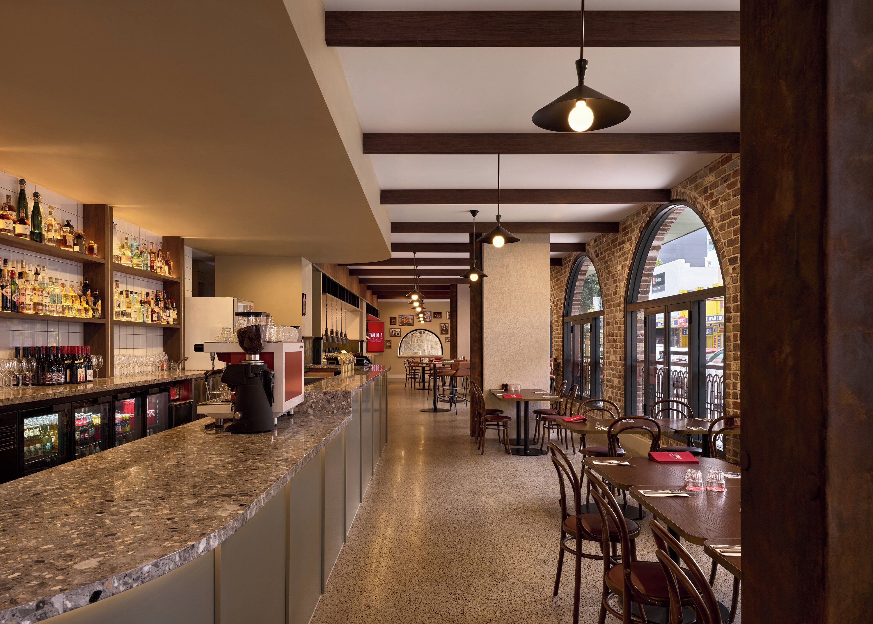 A bar area with a long counter, shelves with bottles, and a coffee machine. There are tables and chairs in the dining area with large windows.