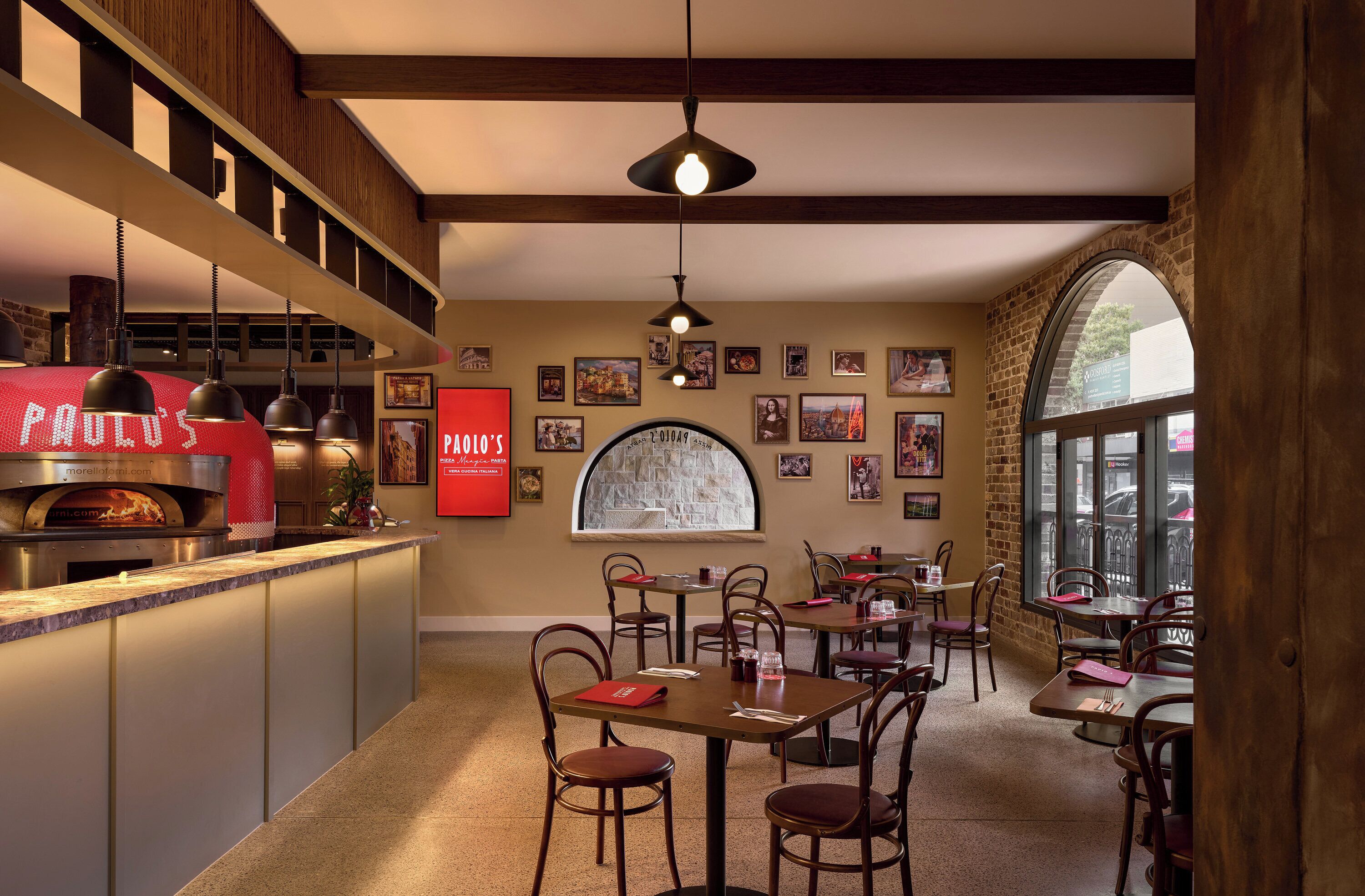 A restaurant interior with a brick wall, wooden ceiling beams, a central arched window, and a pizza oven. Tables and chairs are arranged for dining.