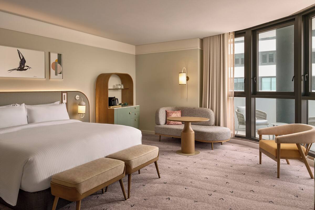 A hotel room with a large bed, a desk, two chairs, a bench, and a large window offering a view of the city.