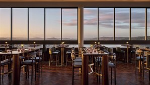 Un restaurante con varias mesas y sillas, amplios ventanales que ofrecen vistas a la ciudad y a las montañas, y un suelo de madera.