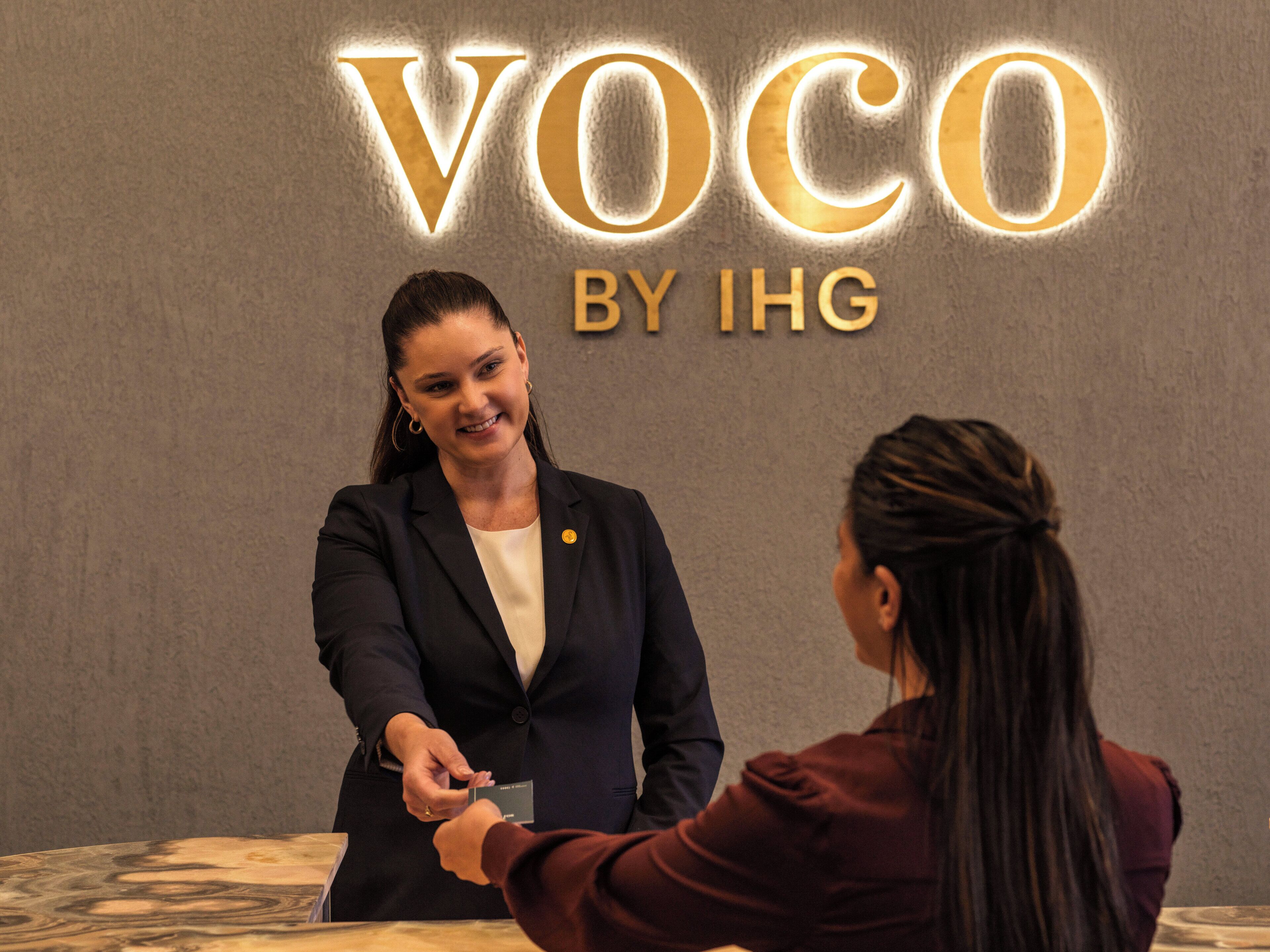 Une personne remet une carte à une autre dans un cadre professionnel, avec une enseigne « Voco by IHG » en arrière-plan.