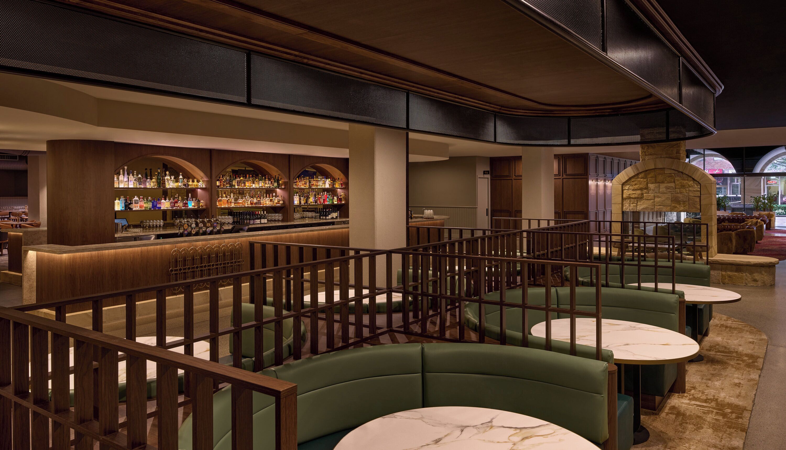 A bar area with a high ceiling, wooden beams, and a stone fireplace. There are green cushioned seating areas with marble-topped tables.