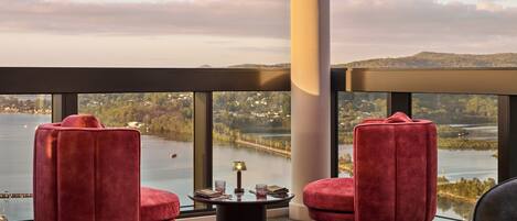 Una habitación moderna con dos sillones de terciopelo rojo, una mesa redonda y vistas a un lago y un paisaje lejano.