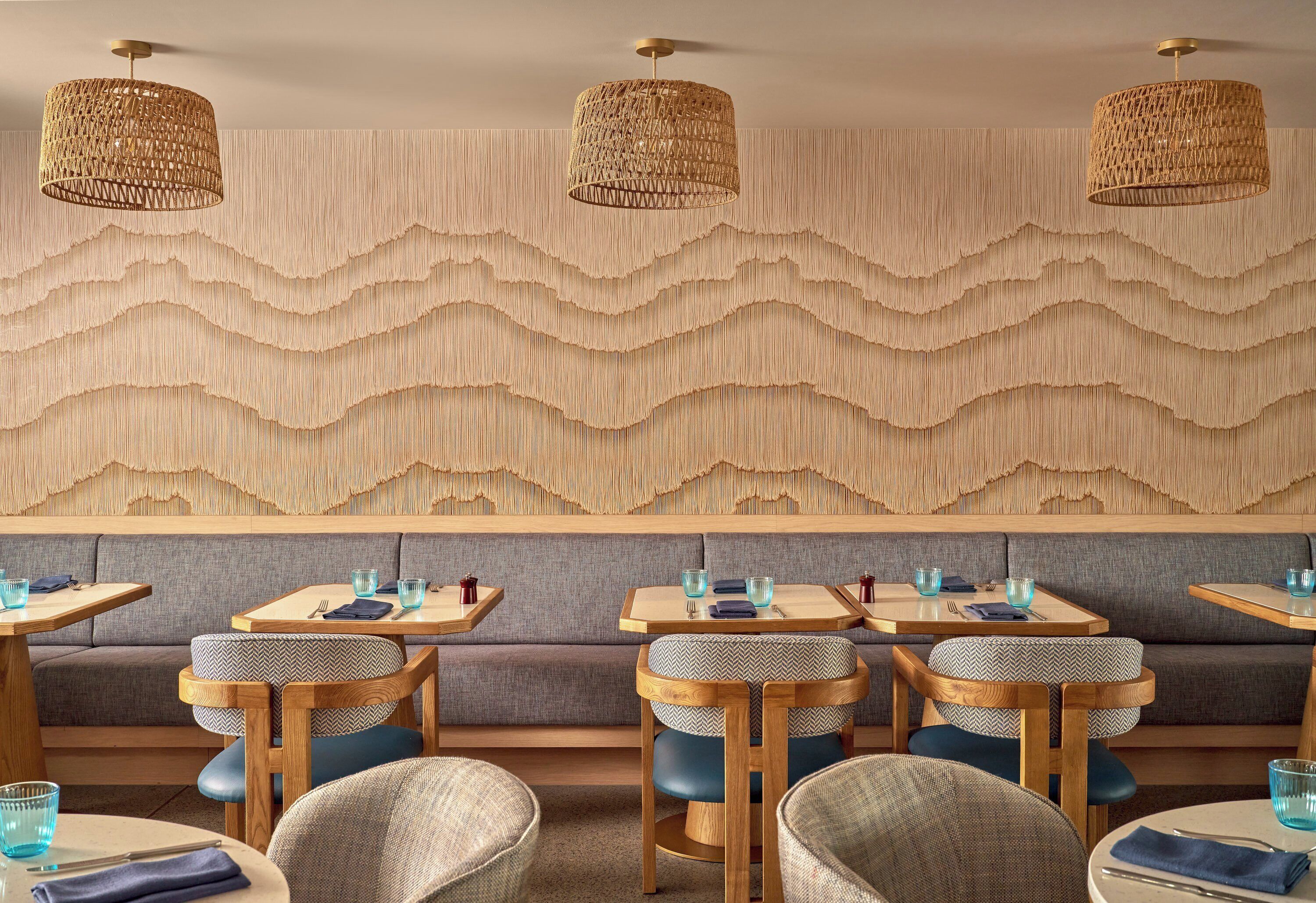Interior de restaurante con mesas y sillas de madera, pared con textura y luminarias colgantes tejidas.