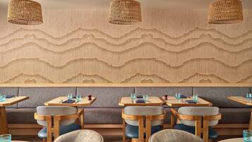 Interior de restaurante con mesas y sillas de madera, pared con textura y luminarias colgantes tejidas.