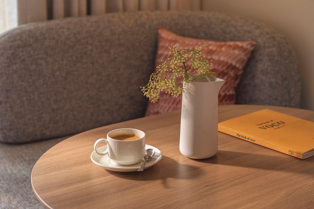 A round wooden table with a white coffee cup and saucer, a white vase with a plant, and a book titled "The Book of Voco".
