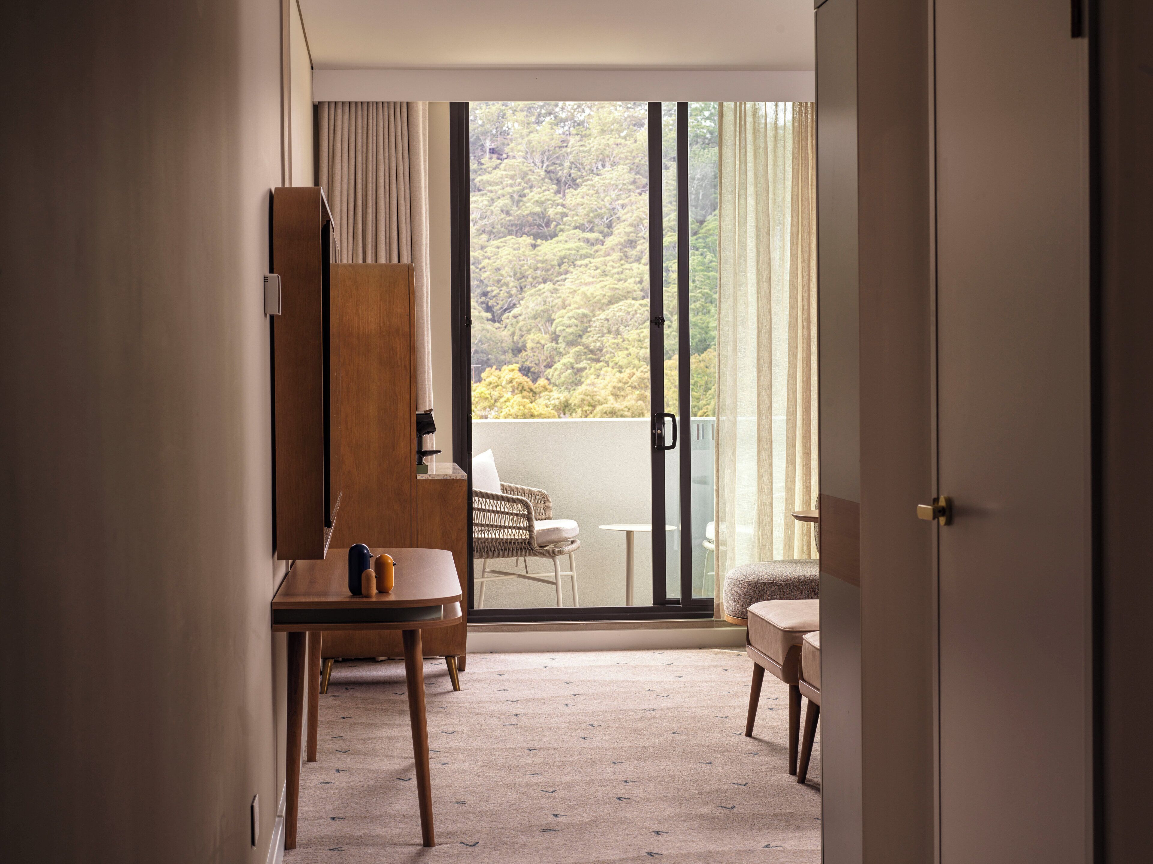 A room with a large sliding glass door leading to a balcony with a view of trees. There is a wooden chair and a small table with a cup on it.