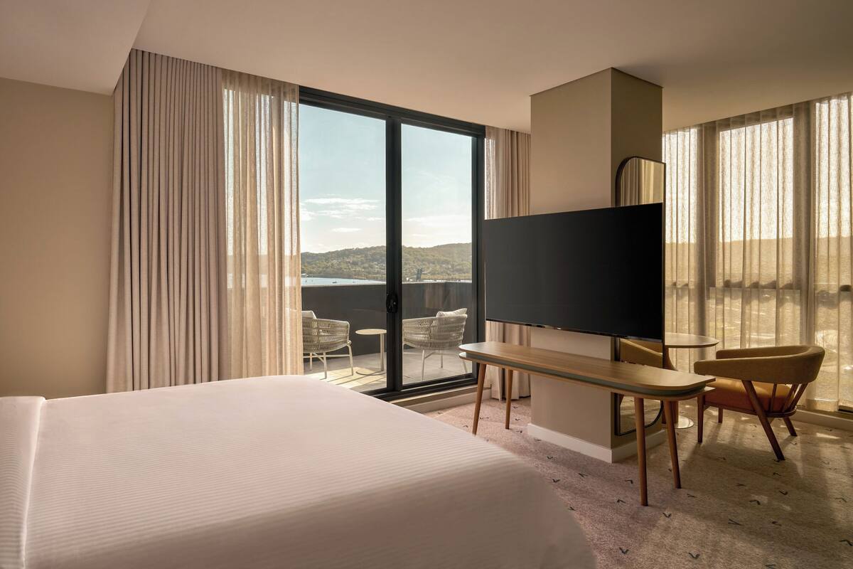 A hotel room with a large bed, a TV, a desk, and a chair. There is a view of the outdoors through the window.