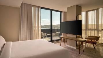A hotel room with a large bed, a TV, a desk, and a chair. There is a view of the outdoors through the window.