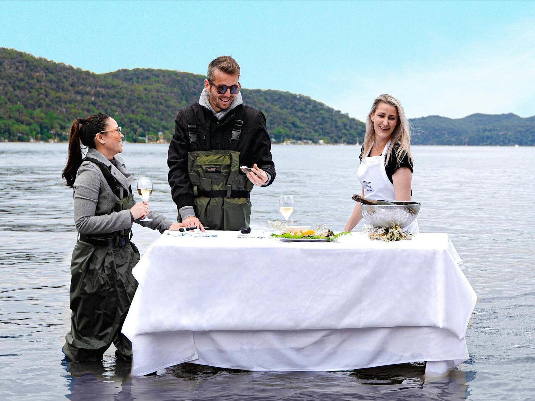 Three people dining at a table on a lakeside with a view of hills in the background.
