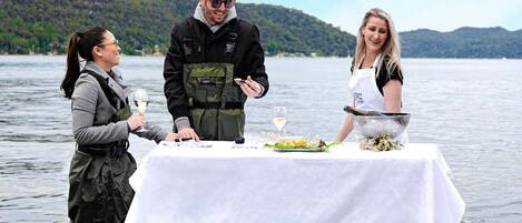 Tres personas cenando en una mesa junto a un lago, con vistas a colinas al fondo.