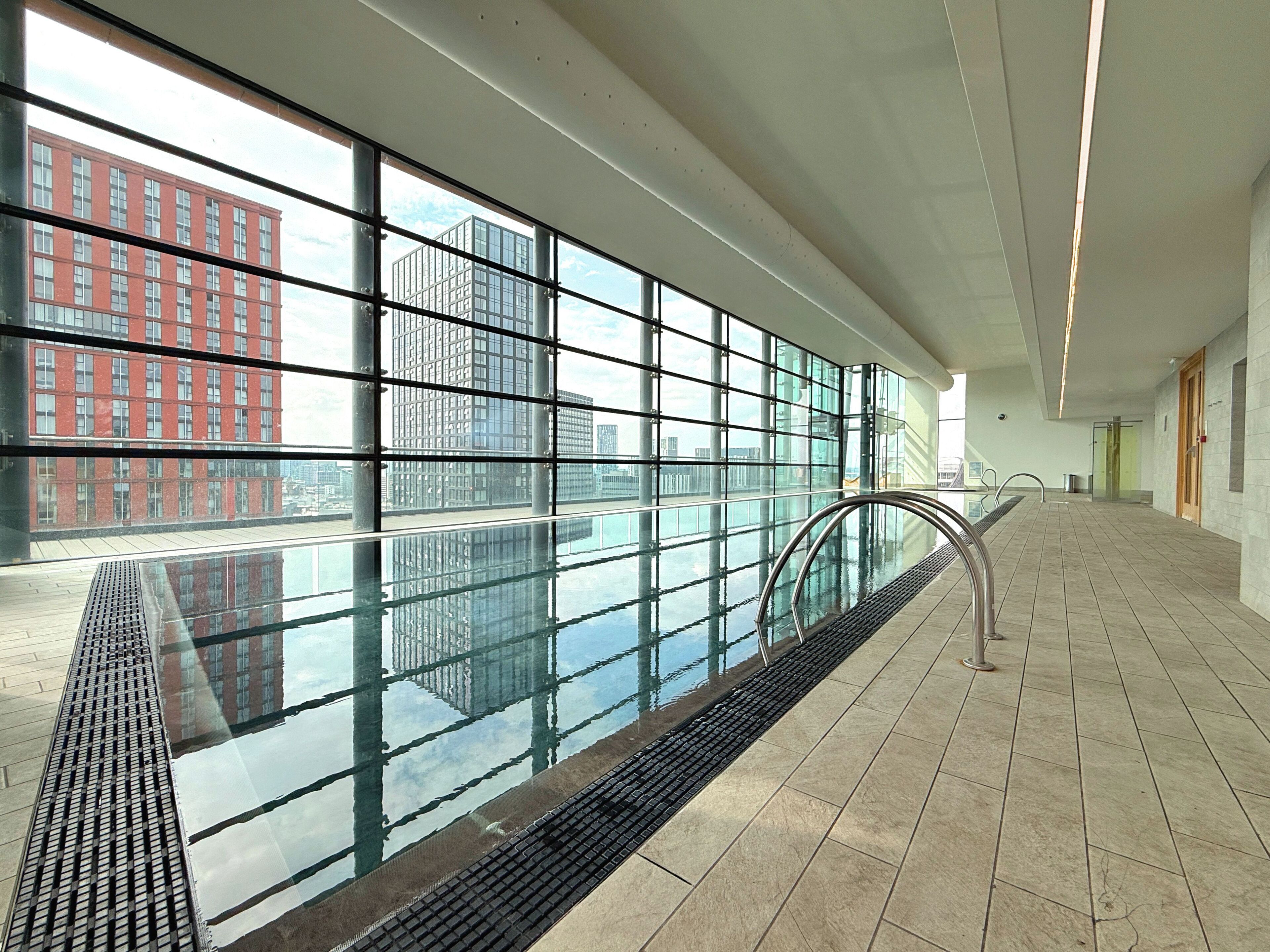 Una piscina interior moderna con paredes de vidrio transparente, un pasamanos de metal y suelo de baldosas.
