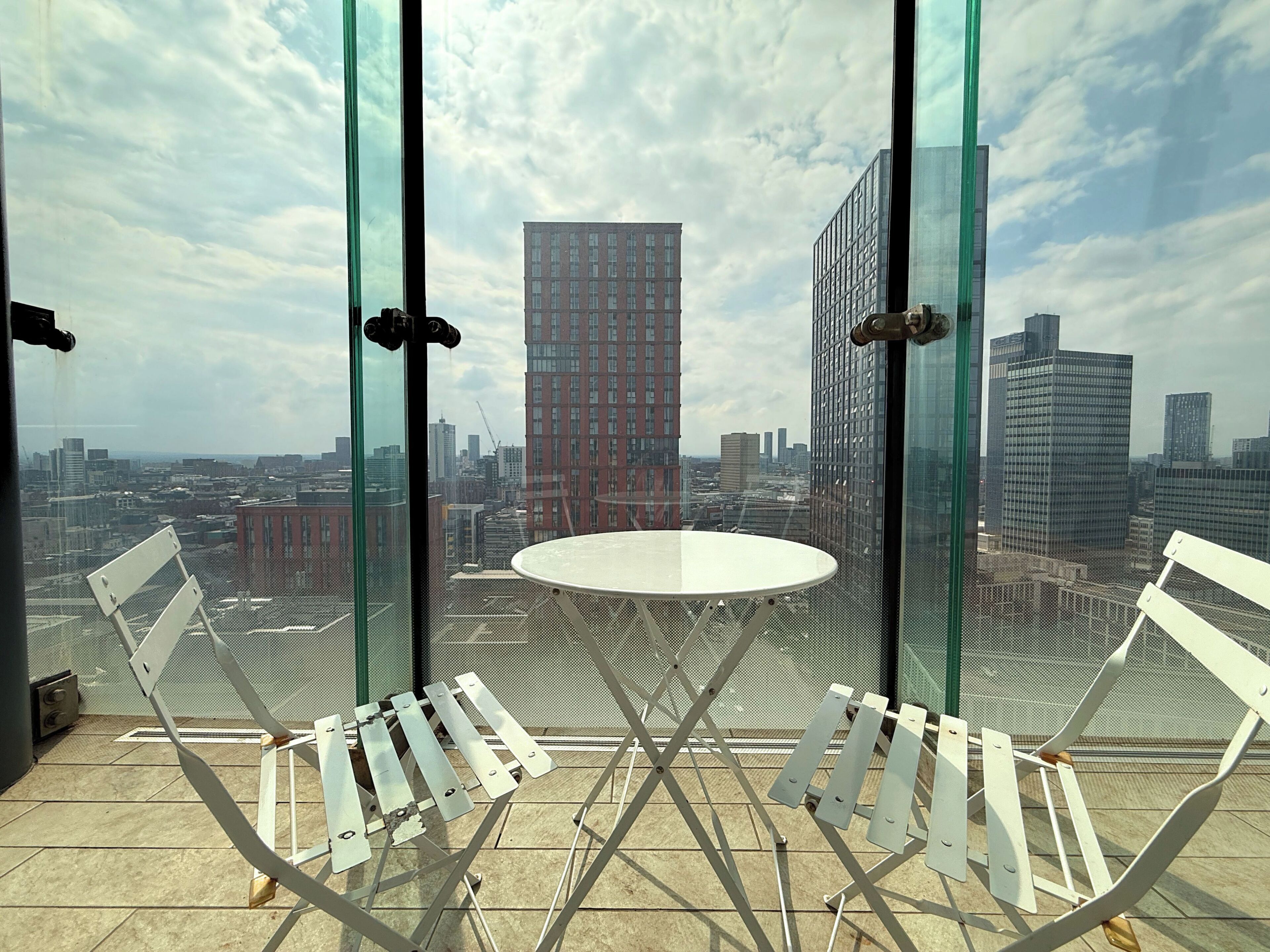 A balcony with a table and chairs, overlooking a cityscape with various buildings.