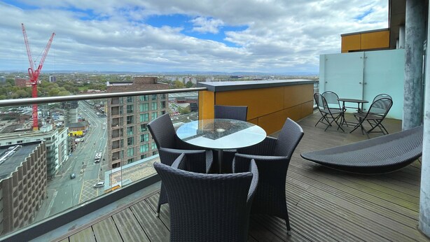 A balcony with wicker furniture, a glass table, and a view of a cityscape including a construction crane. - Modern Luxury in Manchester (Manchester)