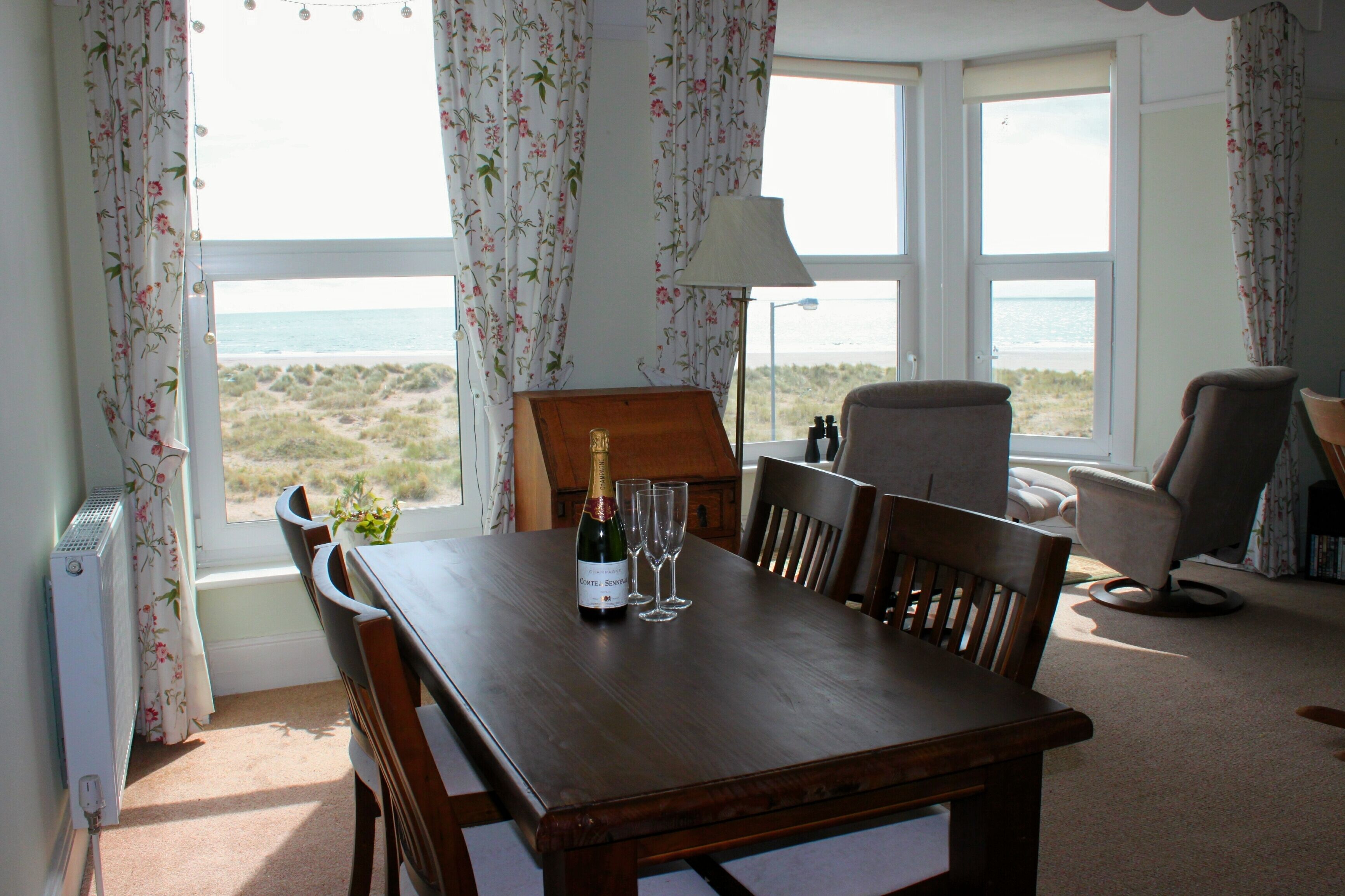 A dining table with a bottle of champagne and two glasses, a lamp, and a view of the beach through large windows.
