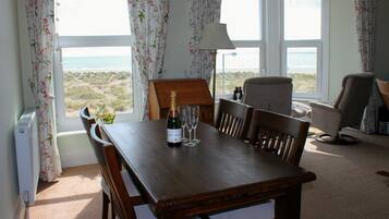 A dining table with a bottle of champagne and two glasses, a lamp, and a view of the beach through large windows.