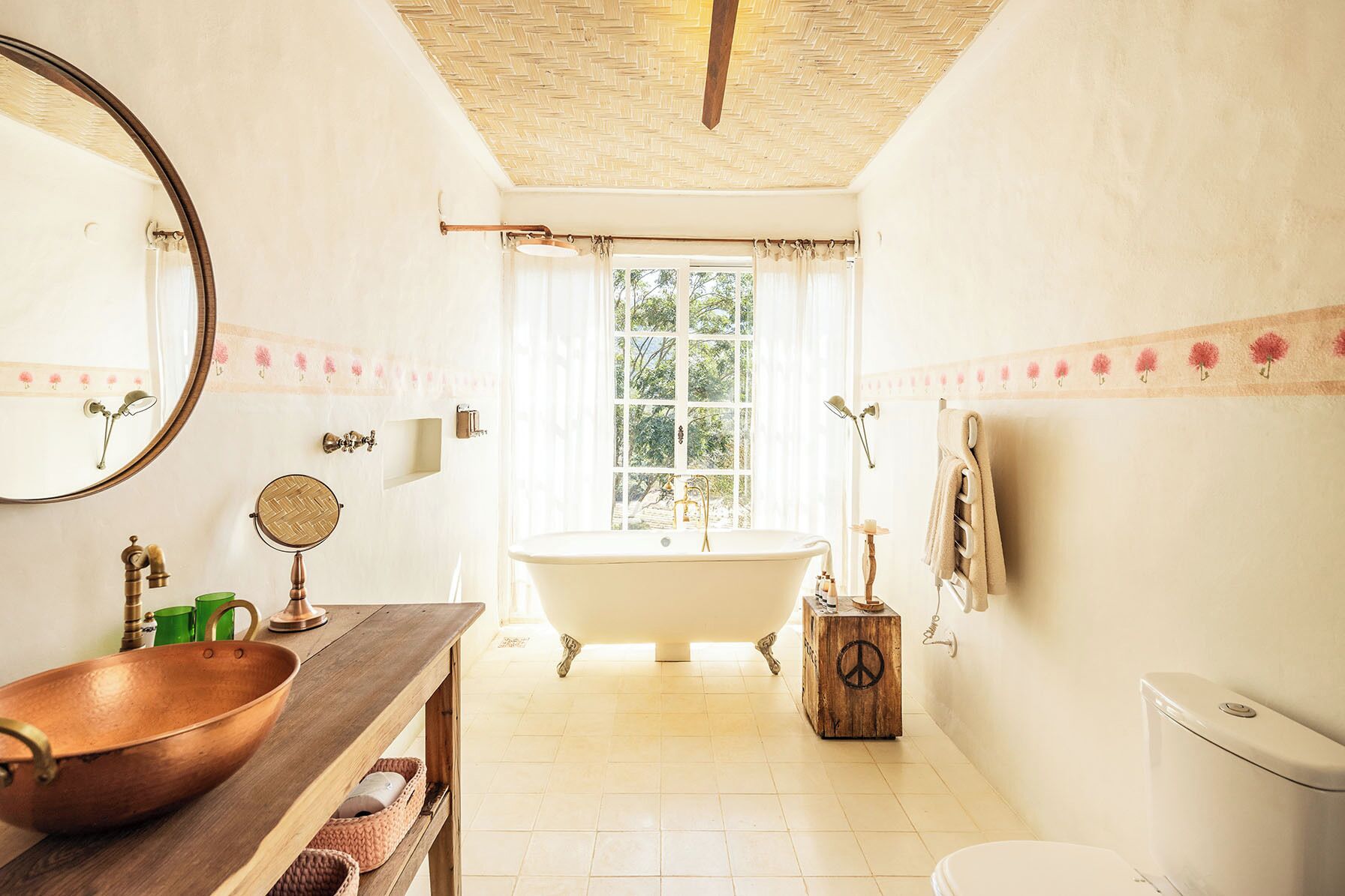 A bathroom with a freestanding bathtub, a wooden vanity with a copper basin, a round mirror, and a large window with curtains.
