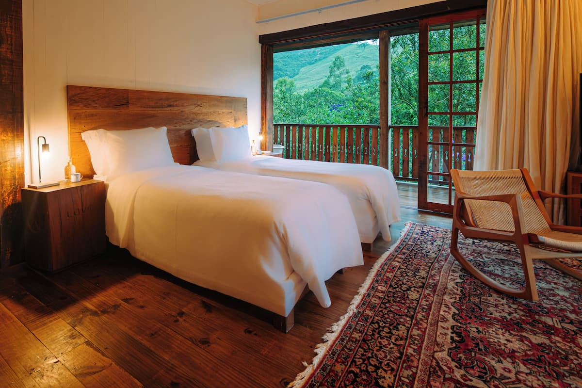 A double bed with a wooden headboard, a nightstand with a lamp, a rocking chair, and a view of greenery through large windows.