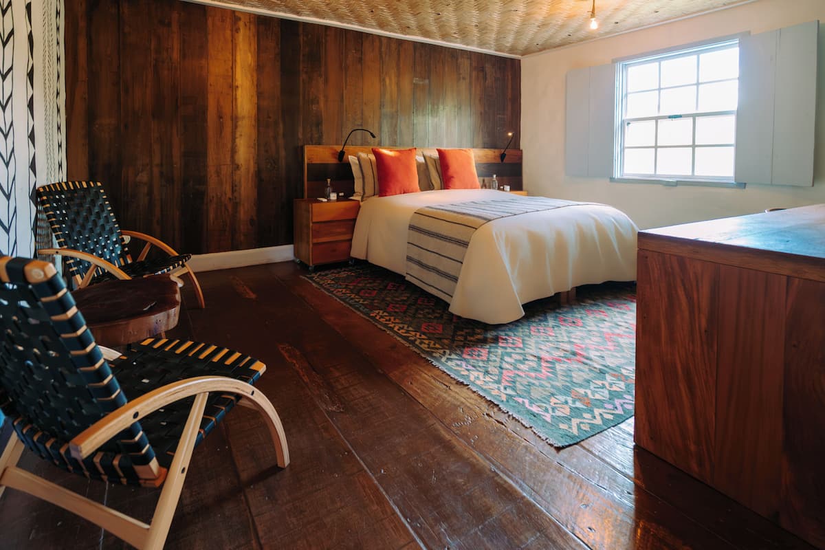 A bedroom with a wooden headboard, a bed with multiple pillows, a nightstand, a chair, and a window.