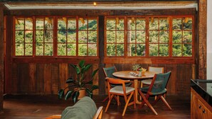 A room with a large window, wooden walls, a round table with chairs, and a potted plant. - Ibiti Village (Lima Duarte)