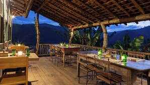 An outdoor dining area with wooden tables and chairs, set against a backdrop of mountains and tropical plants. - Ibiti Village (Lima Duarte)