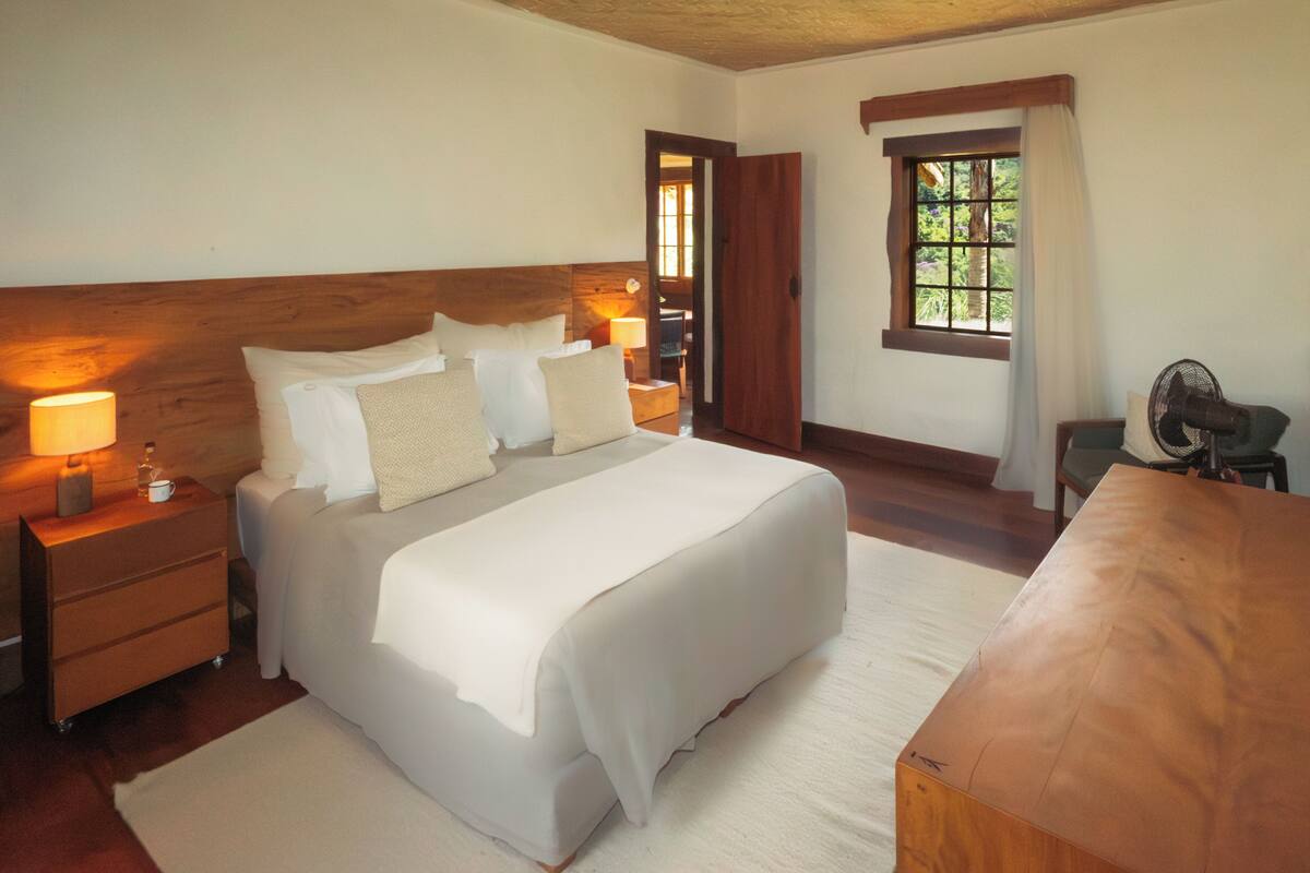 A neatly arranged bedroom with a bed, bedside tables, a nightstand, a wooden headboard, a window with curtains, and a fan.