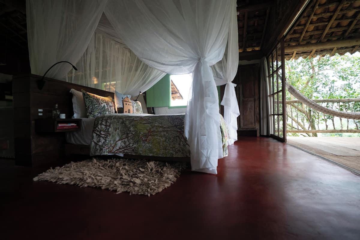 A bedroom with a canopy bed, a bedside table with a lamp, and a view of the outdoors.