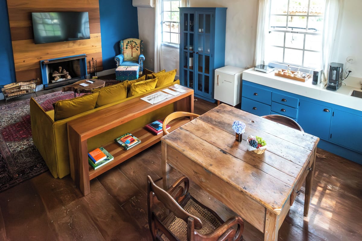 A cozy living room with a wooden sofa, a coffee table, a fireplace, a television mounted on the wall, and a kitchen area with blue cabinets.