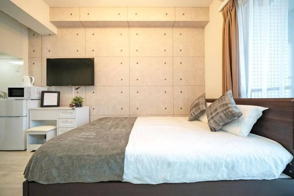 A modern bedroom with a bed, a flat-screen TV mounted on the wall, a desk with a chair, and a window with curtains.