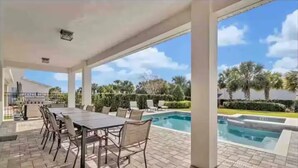 A covered outdoor area with a pool, a dining table with chairs, and a view of palm trees and a clear sky.