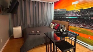 A room with a bed, a table, a TV mounted on the wall, and a mural of a baseball stadium. - Downtown Boston Famous Nightlife Central Location (Boston)