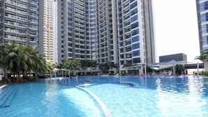 A swimming pool surrounded by tall residential buildings, palm trees, and a covered area with umbrellas. - Atlantis Residence Premium By I Housing (Melaka)