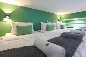 Quarto de hotel com duas camas, paredes verdes, roupa de cama branca e cabeceira.