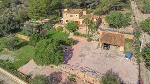 Una casa de piedra de dos pisos con techo de tejas, rodeada de un jardín con piscina y un coche pequeño aparcado afuera.