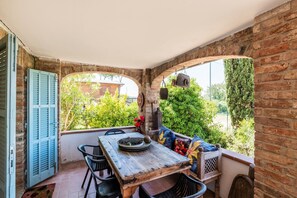 Eine gemütliche Terrasse mit Holztisch und -stühlen, einer gepolsterten Bank und Blick auf Grünflächen und eine Ziegelwand.