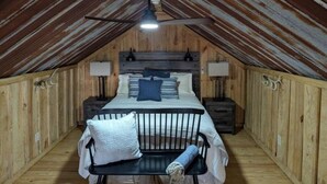 A cozy attic bedroom with a wooden ceiling, a bed with pillows, a bench, and two bedside tables with lamps. - Hidden Oaks Lavish Cabin #2 (Abbeville)