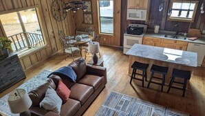 A cozy cabin interior with a wooden dining area, a sofa, a kitchen with a bar, and a large window. - Hidden Oaks Lavish Cabin #2 (Abbeville)