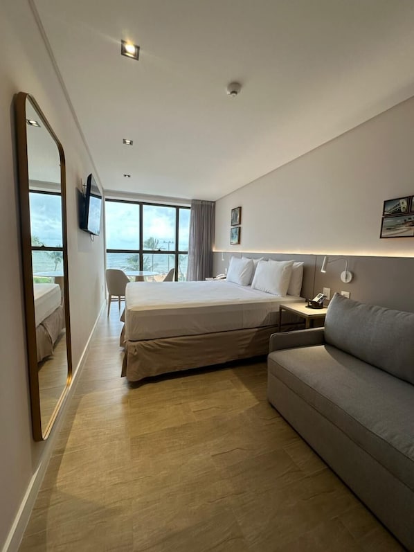 Quarto de hotel com cama, sofá, escrivaninha, cadeira e vista para o oceano.