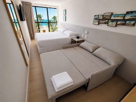 Una camera d'albergo con un letto grande, un letto più piccolo, un divano, un comodino e vista sulla spiaggia.