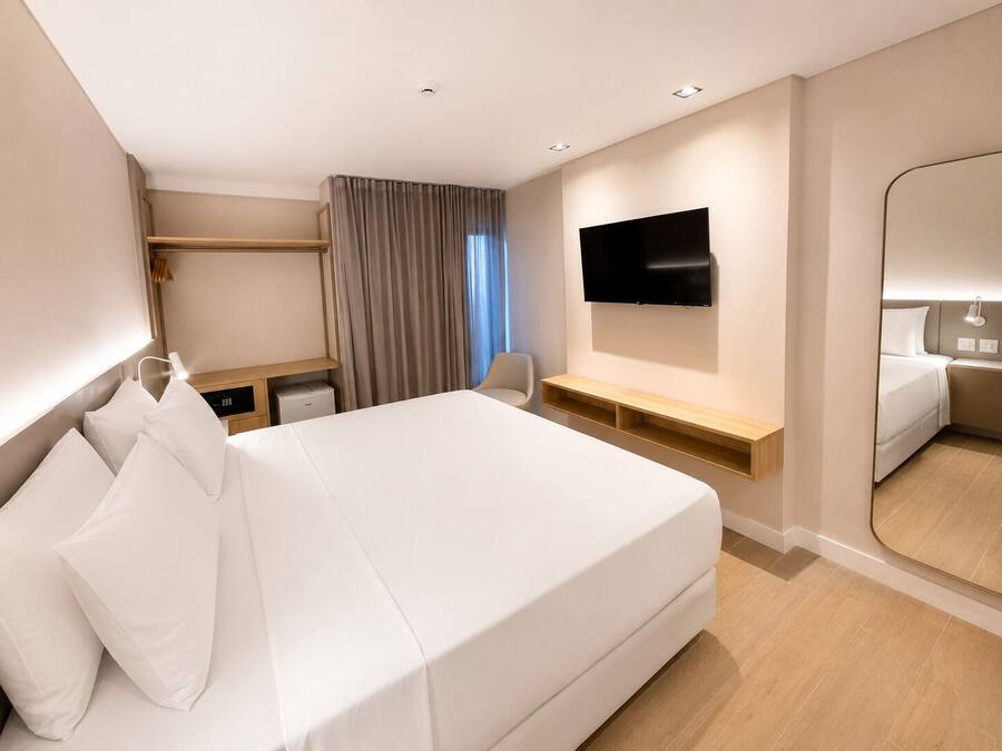 A modern hotel room with a large bed, a flat-screen TV mounted on the wall, a bedside table, a desk with a chair, and a large mirror.