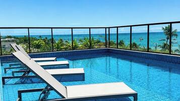 A clear blue pool with lounge chairs, a tiled deck, and a view of the ocean.