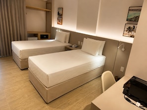 Quarto de hotel com duas camas, uma escrivaninha, uma cadeira, um telefone e uma pequena mesa com luminária.