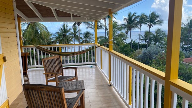 Ein überdachter Balkon mit Holzstühlen und einer Hängematte, mit Blick auf eine tropische Landschaft mit Palmen und dem Meer.