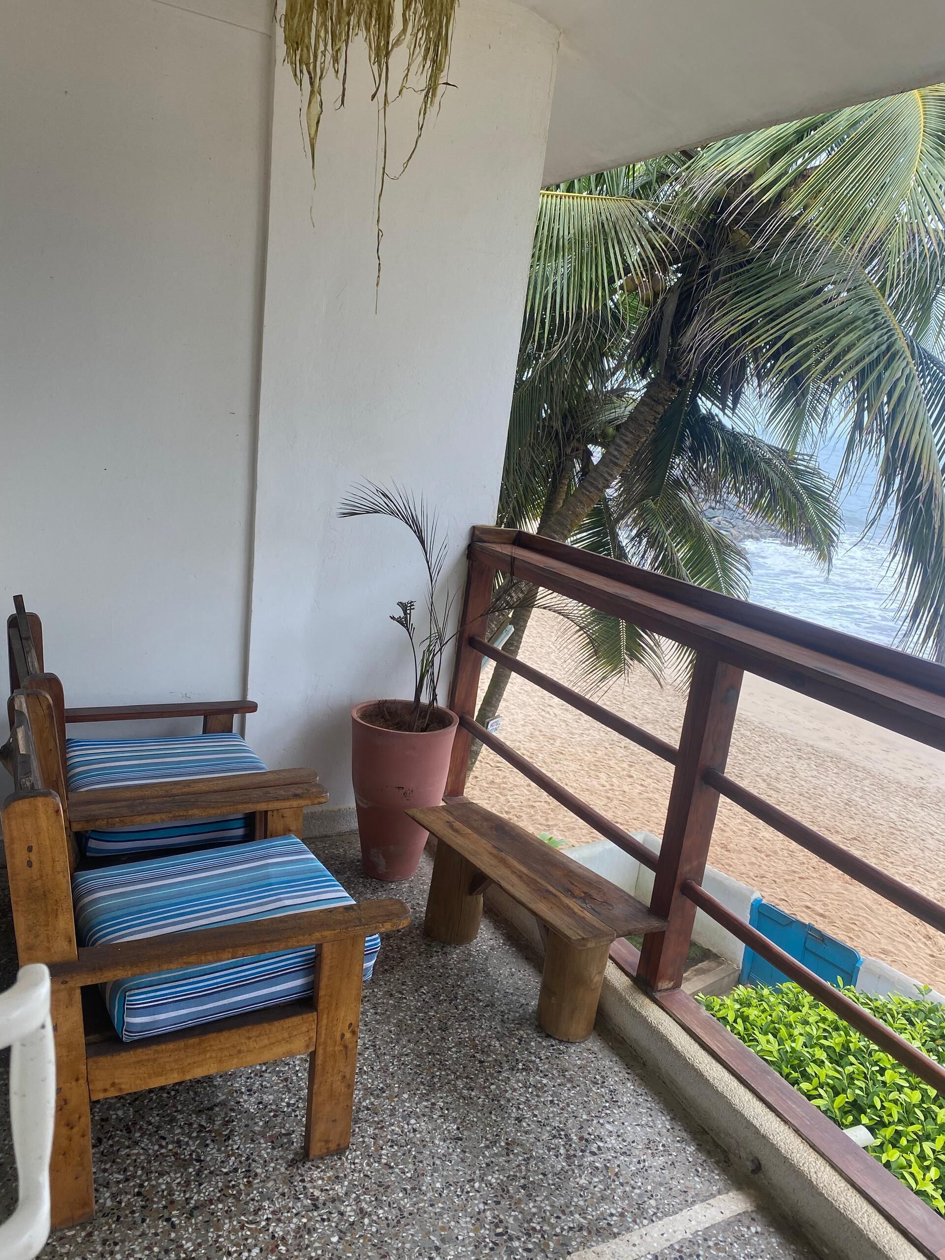 A wooden bench and chairs with striped cushions on a balcony with a view of the beach and palm trees.
