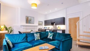Uma sala de estar moderna com um sofá azul-turquesa, mesa de centro de madeira e uma cozinha ao fundo.