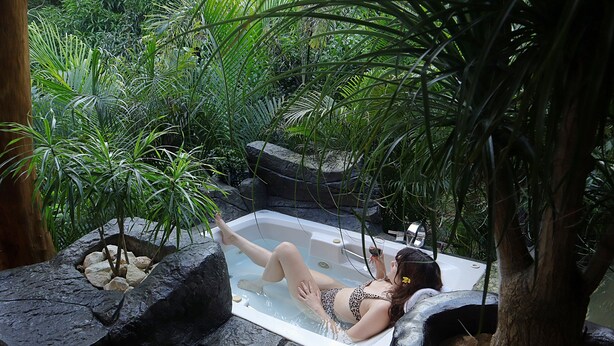 Une femme se détend dans un bain à remous, entourée d’une végétation luxuriante et de rochers.