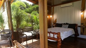 Une terrasse en bois avec des meubles en osier, un lit avec une tête de lit en bois et une vue sur une végétation luxuriante.