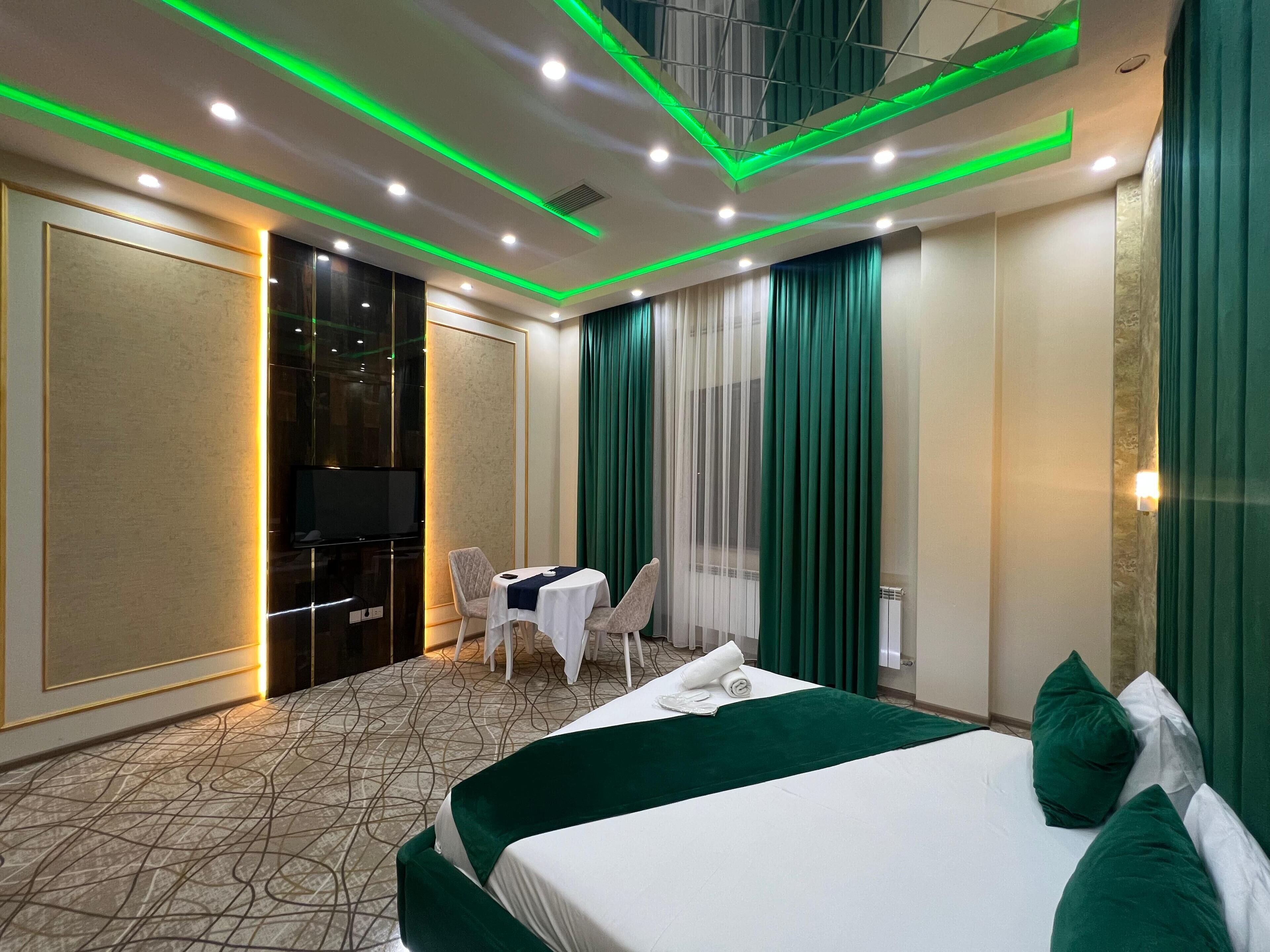 A hotel room with a bed, a desk, and a chair. There is a television on the left side of the room. The room has green curtains and a patterned carpet.