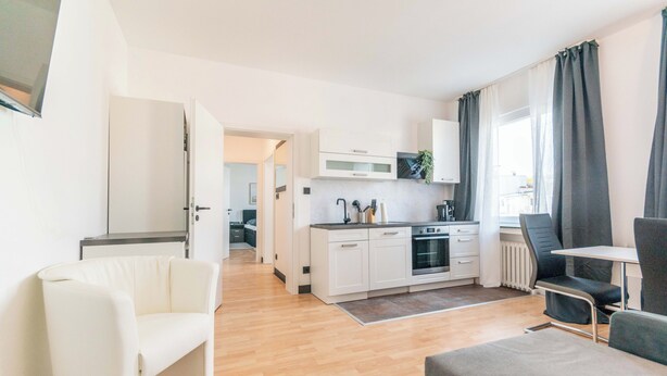 Una sala de estar moderna y bien iluminada, con sofá, mesa de comedor y área de cocina con armarios blancos y horno empotrado.