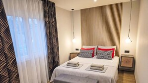 Un dormitorio moderno con una cama grande, dos mesitas de noche, un banco y una ventana con cortinas translúcidas.
